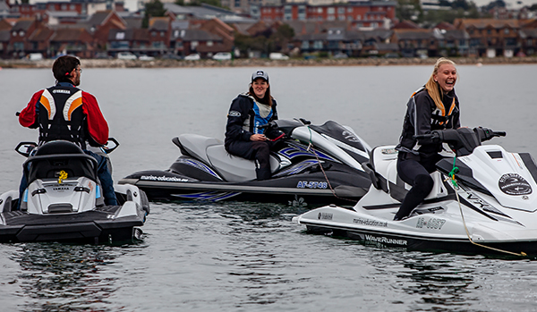 Two women and a man on personal watercraft. Female RYA instructor is running a training course. Blonde woman is looking at the camera and laughing. Houses and cars are visible ashore in the background.