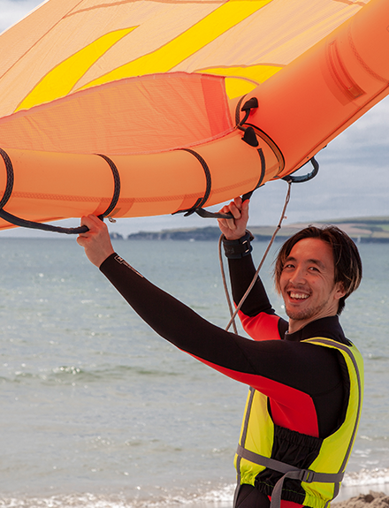 Man holding orange inflated wing on beach. Smiling at camera. Learning wing control before going afloat on RYA Learn to Wingsurf course.