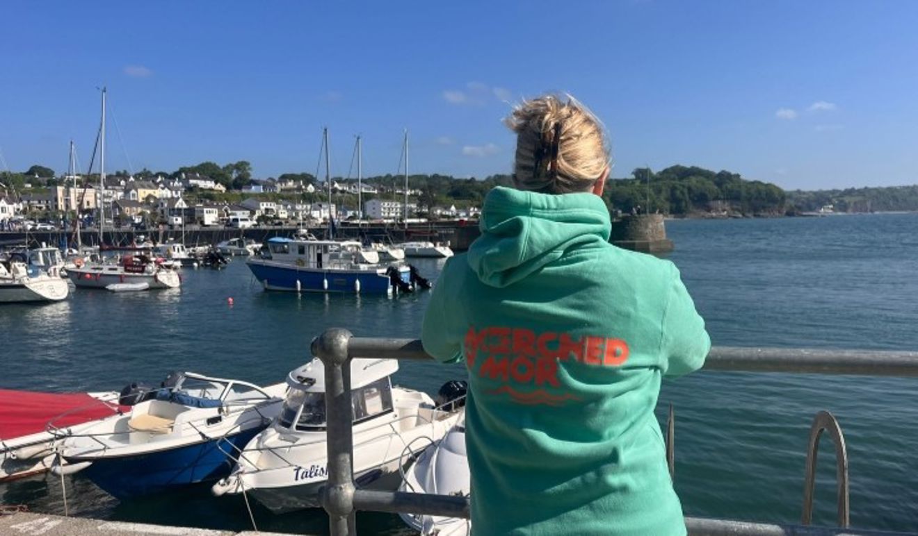 Woman in a Merched Y Mor branded hoody looking out over a harbour on a sunny day with blue skies.