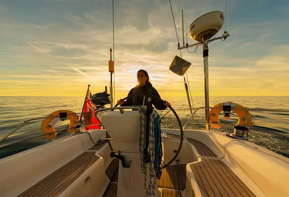A sailor helming (steering) a yacht as the sun sets in the background