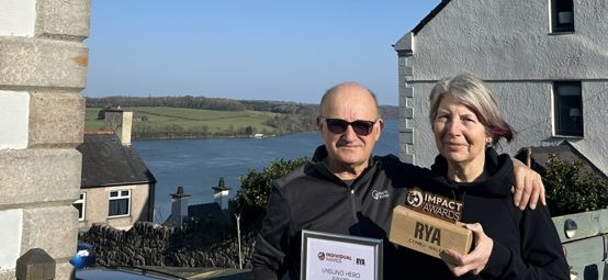 Moley and Mandy Morsley with their Welsh Sailing Impact Award on a sunny day in Port Dinorwic with houses and the sea in the background.