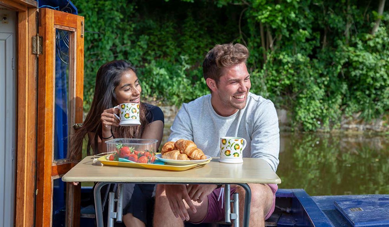 Man and a woman sat on a narrowboat, enjoying a drink and some food