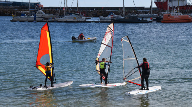 Long shot of three youths on windsurfs with boats in the background