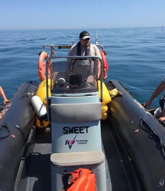 A man sat on a Barcelona Watersports rigid inflatable boat (RIB) with people wearing lifejackets on jet skis on either side of it on a beautiful sunny day