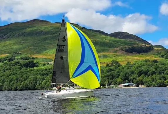 A small sailing boat with blue and yellow spinnaker sailing on the water with lush green hills in the background