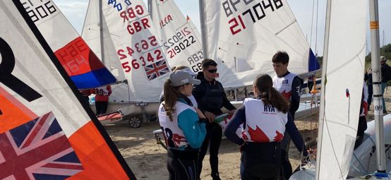 Young sailors chatting in a group alongside rigged boats on the beach at the Welsh Youth & Junior Championships, Plas Heli, 2025.
