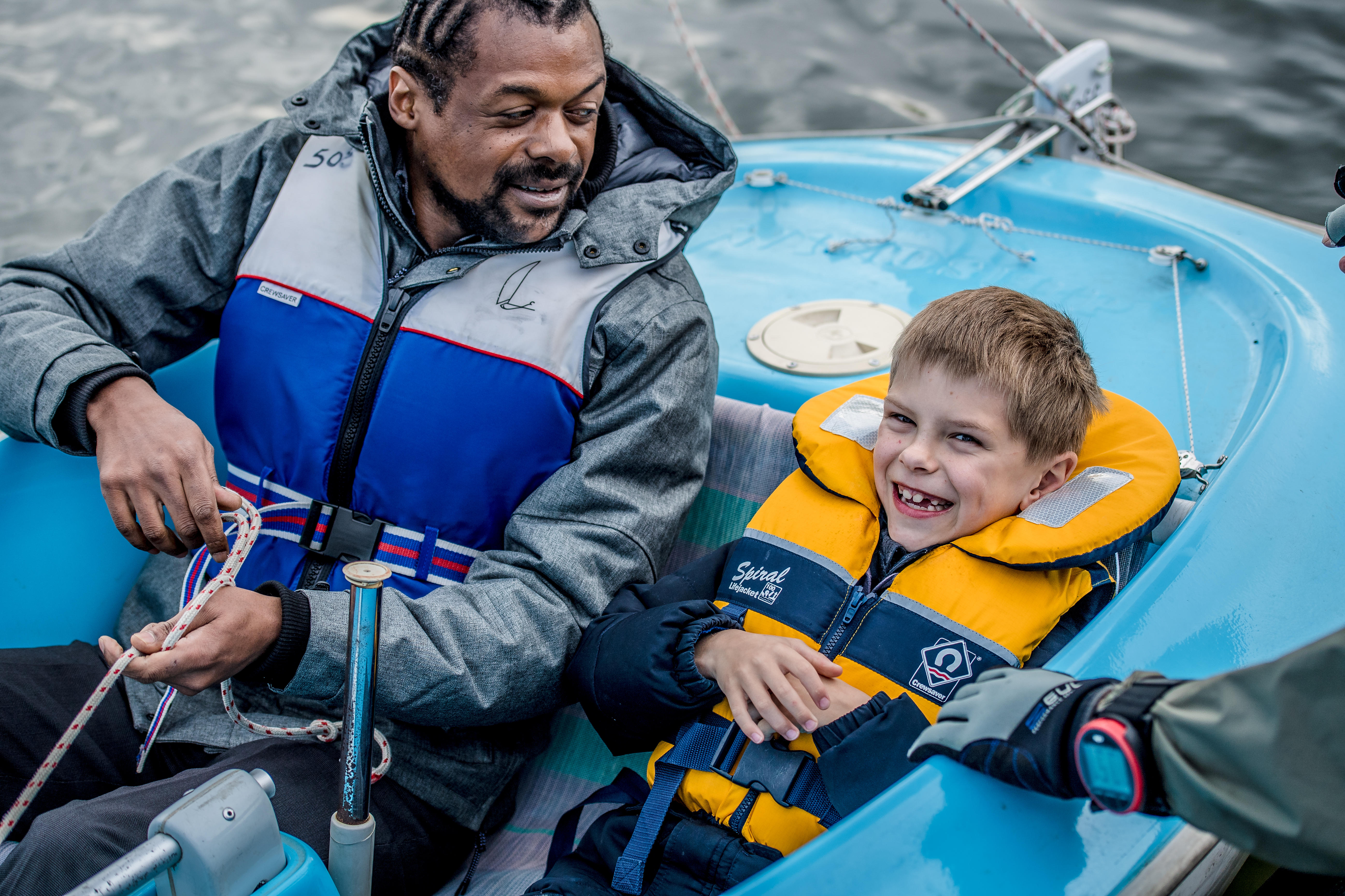 man and a child sailing a dinghy, man is looking at the child, and the child is smiling and looks excited