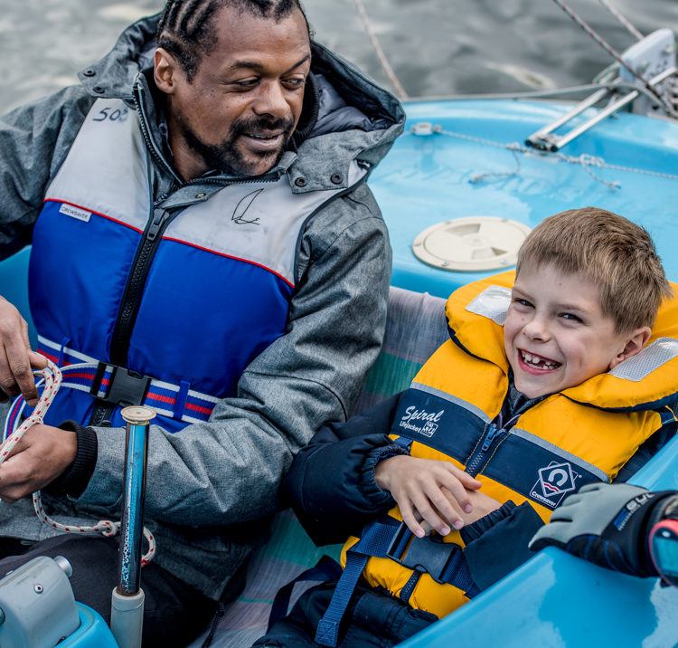 man and a child sailing a dinghy, man is looking at the child, and the child is smiling and looks excited