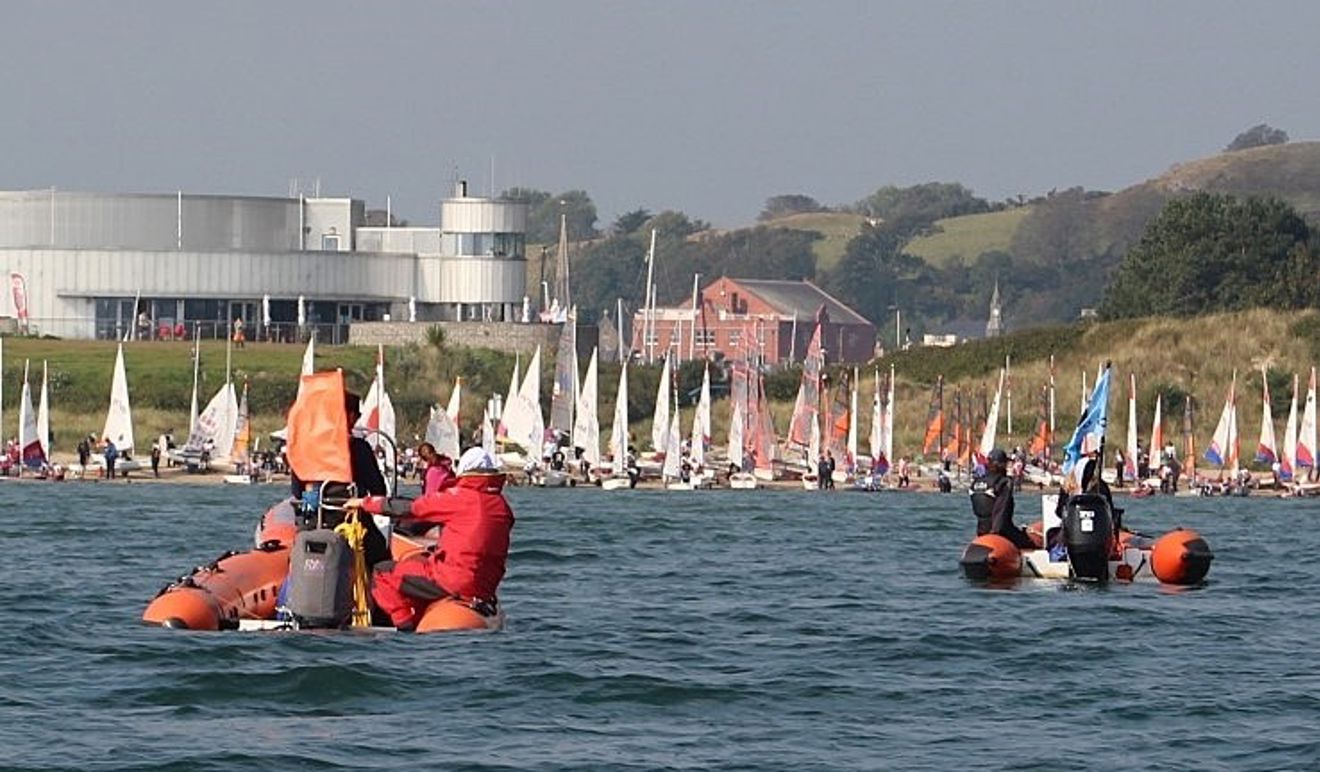 View of the Welsh Y&J Championships from the sea with two powerboats on the water waiting for 83 boats to launch in front of the Plas Heli venue, which is shiny silver in the sunshine.