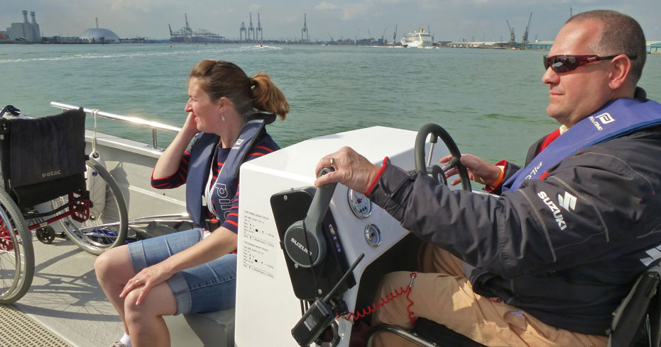 Gentleman in a wheelchair at the controls of a Powerboat, with a lady sat in front of him looking out at the ocean.