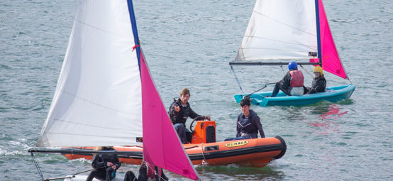 Long shot of sailing dinghys and students being taught by instructors in a powerboat