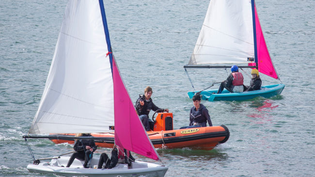 Long shot of sailing dinghys and students being taught by instructors in a powerboat