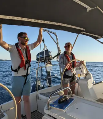 A man with his arms held high, holding the canopy of a sailing yacht while a lady helms (steers) the boat and on a sunny day with perfect blue sky with Yacht Sail Training School, Brac Island, Croatia