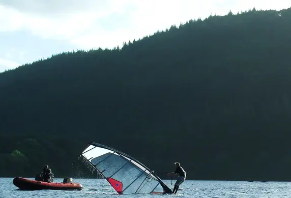 A person practicing pulling up a windsurf sail with a safety boat keeps watch