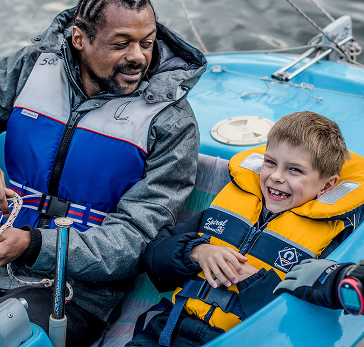 man and a child sailing a dinghy, man is looking at the child, and the child is smiling and looks excited