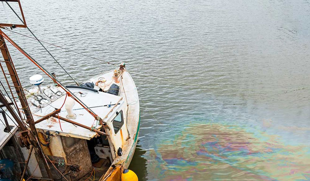 An Oil Slick In The Water Next To An Old Dilapidated Fishing Boat. The Slick Is Vibrant And Colourful. A Dock Is Visible.