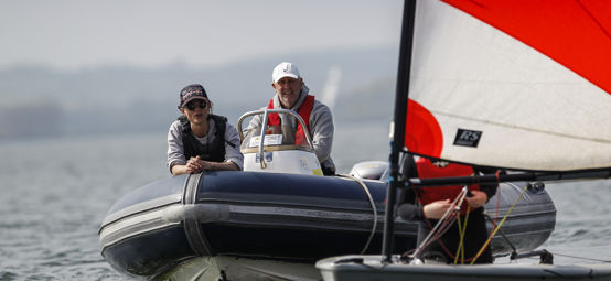 Long shot of man and woman on dinghy with sailboat blurred out in the foreground