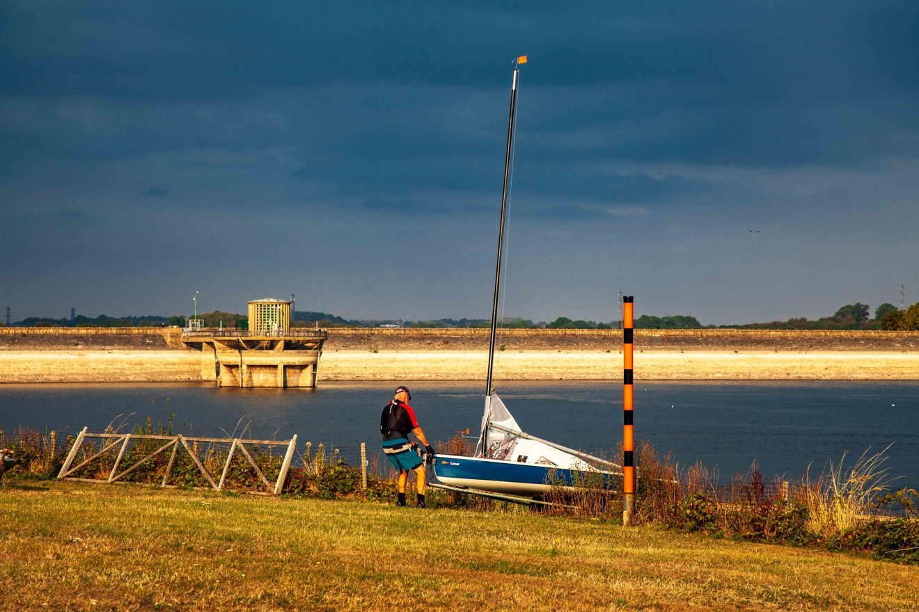Long shot image of a person with a sailing dinghy on the grassy bank beside a reservoir.