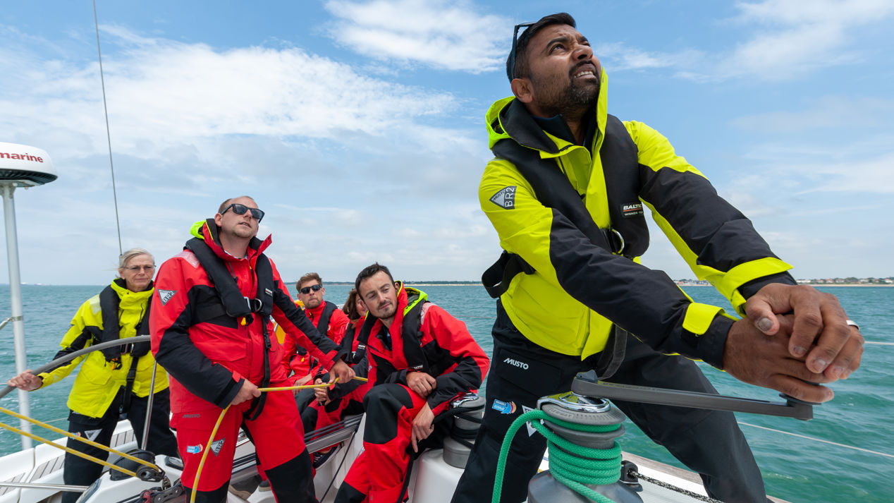 Mid-wide shot of crew operating sailboat on calm sea under partly cloudy sky