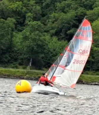 A small sailing boat with clear and red striped sailing heeling (leaning over) as it rounds a mark on the water