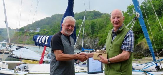 Cardigan Bay Watersports principal Crawford McLeary presents an RYA Cymru Wales Impact Award to Stephen Wood, onshore with keelboat in background.