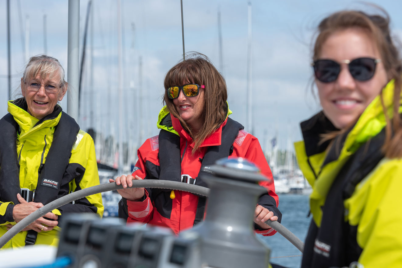 Three women at the stern of a yacht, one at the wheel, all smiling