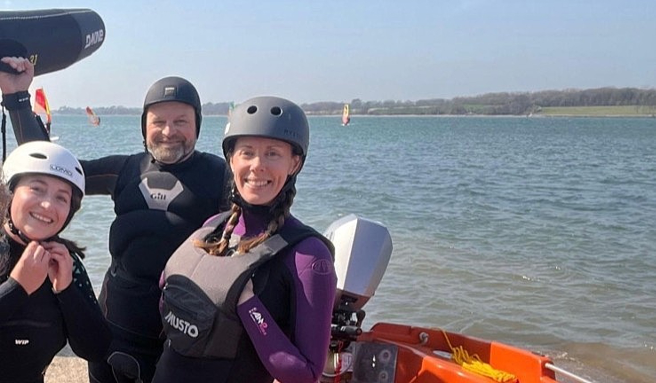 Three members of the RYA CW team - Hatty Morsley, James Stuart and Sarah McGovern - smiling for the camera while wearing helmets, boyancy aids and wetsuits and holding aloft a board during a 'have a go at winging' session.