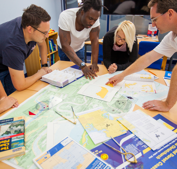 Group of adults undertaking navigation chart work in classroom, pilot guide, course plotter, dividers