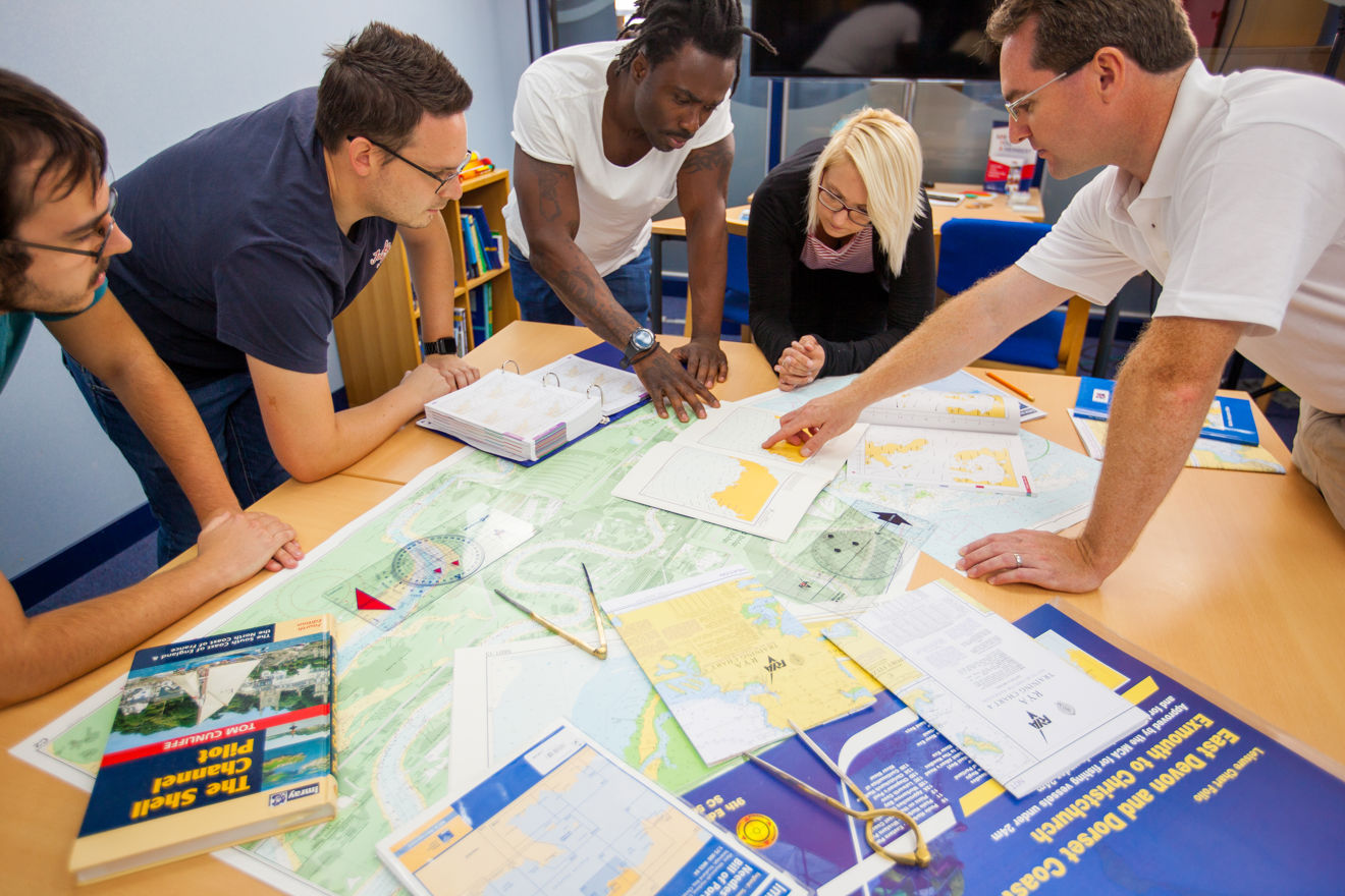 Group of adults undertaking navigation chart work in classroom, pilot guide, course plotter, dividers