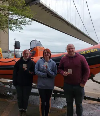 Three people holding certificates in front of Humber Rescue Service rigid inflatable boat (RIB) next to the Humber Bridge