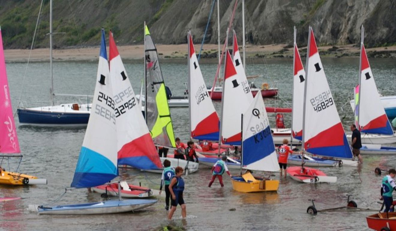 Lots of junior boats with colourful sails and young sailors launching off the beach in OnBoard bibs at Cardigan Bay Watersports.