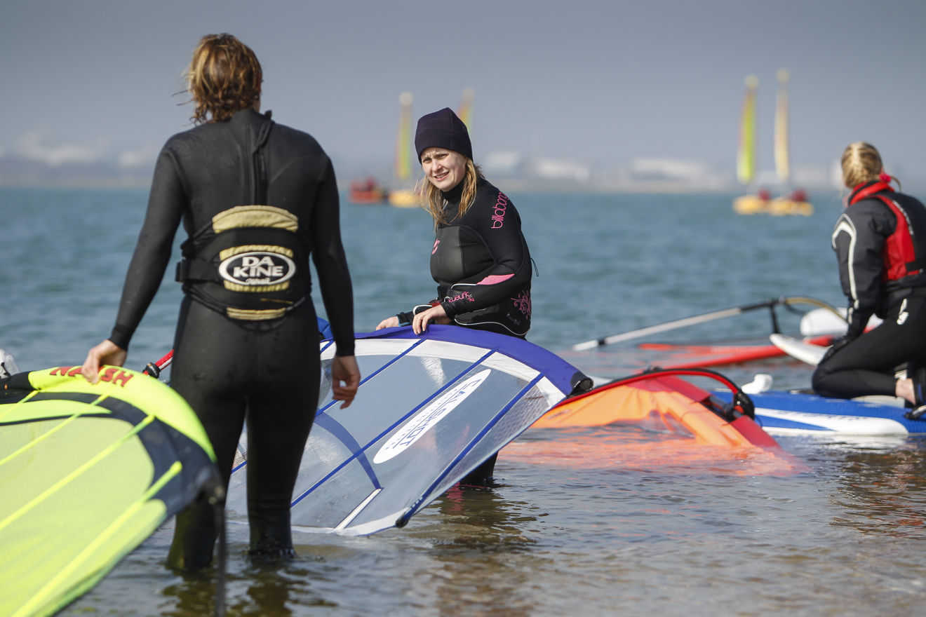 two women with windsurfer equipment in shallow waters talking