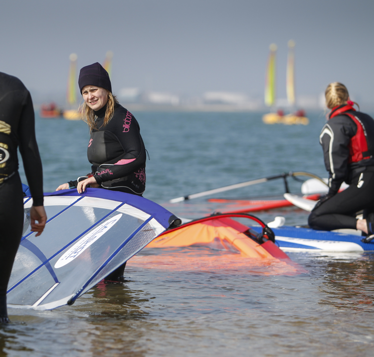 two women with windsurfer equipment in shallow waters talking
