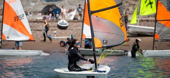 A view from the water of a girl launching from the beach at Dale for a Dragon Series event wearing sunglasses and sailing an RYA Tera with an orange sail, with other children also getting their boats ready to launch.