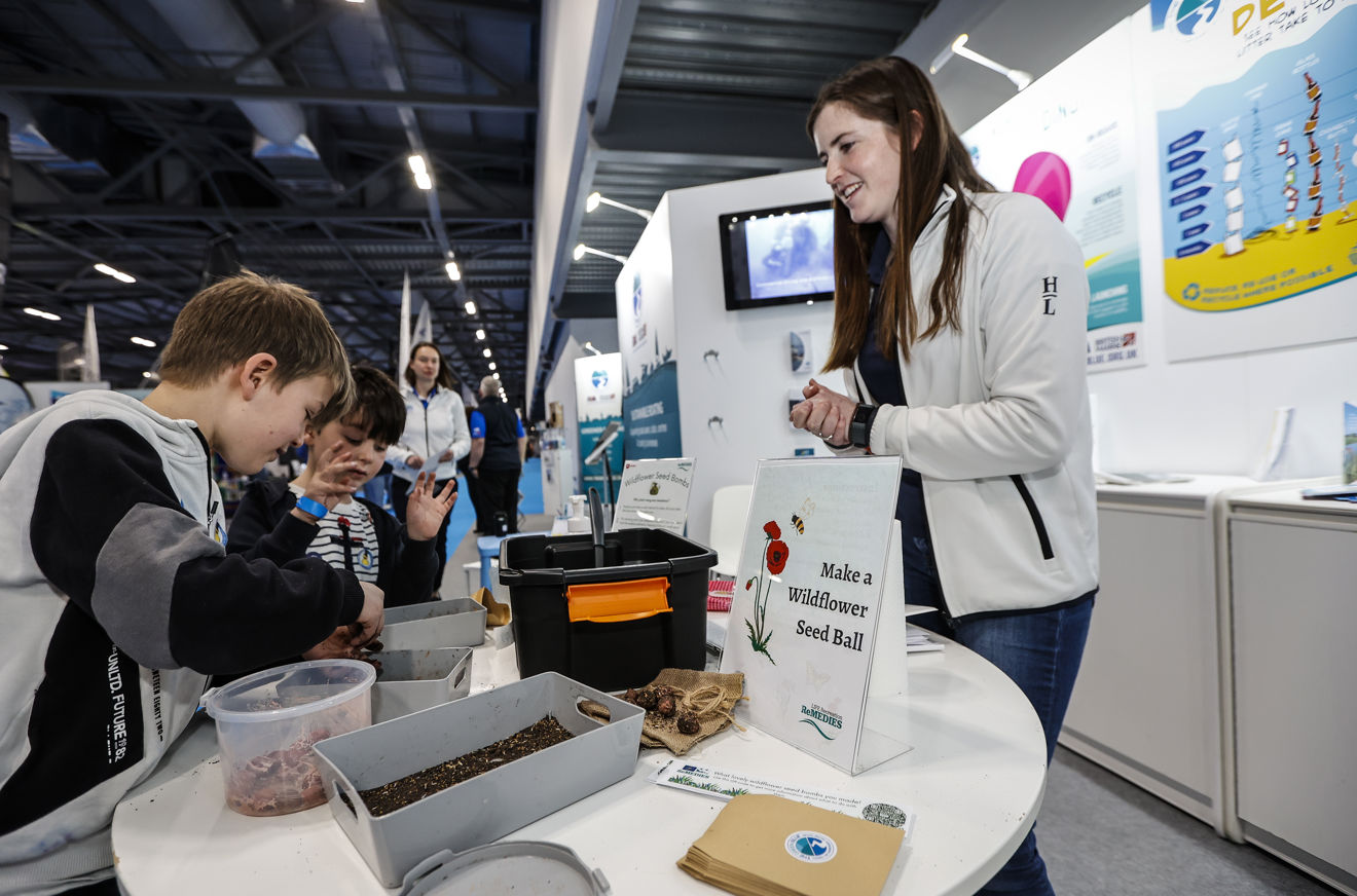 two children sitting at a table at a show, looking at wildflower seeds - woman is standing in front of them telling them about the seeds