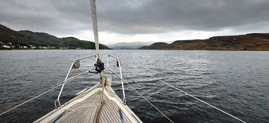 Sloop rigged modern yacht with wooden teak deck sailing on a cloudy day. A view from the deck to the bow. Panoramic view of the rocky shores of Kyles of Bute from the water. Bute island, Scotland, UK