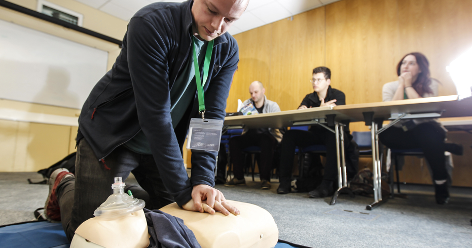 Numerous people in a classroom at a First aid course, one man in knelt on the floor demonstrating CPR on a dummy